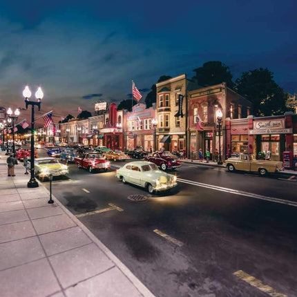 Night view of a Main Street scene with US flags, vintage cars, and illuminated storefronts, showcasing patriotic decor.