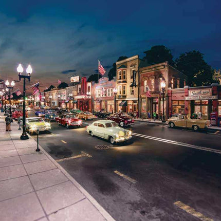 Scenic nighttime view of a vintage-style street with classic cars, illuminated shops, and American flags.