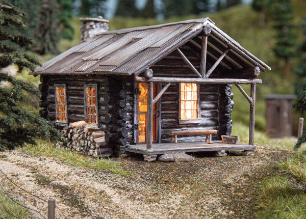 O Scale Cozy Cabin by Woodland Scenics with glowing interior and rustic exterior in a rural setting.